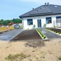 Nowoczesny dom z panelami słonecznymi, świeżo zagospodarowany ogród z wyłożonymi płytami chodnikowymi, przygotowane rabaty z ziemią i posadzone małe tuje wzdłuż ścieżki, w tle mężczyzna i samochód.