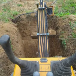 Widok z kabiny minikoparki na wykop w ziemi, z fragmentem osoby w tle. Żółta maszyna z czarnymi elementami sterującymi, otoczona zielenią.