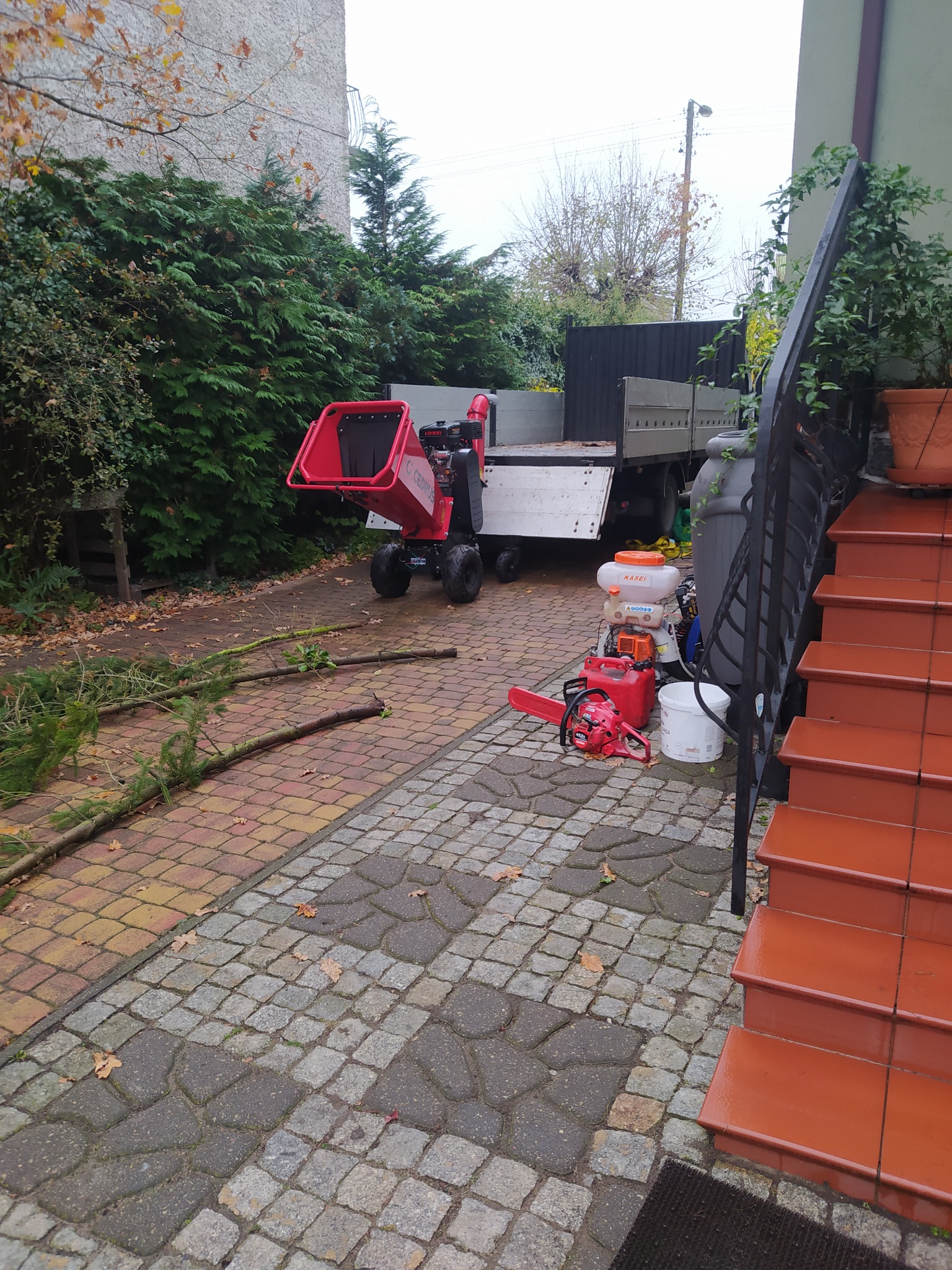 Rozdrabniacz gałęzi na posesji w Bytnicy, obok leżą pocięte gałęzie, piła spalinowa i opryskiwacz, w tle stoi samochód dostawczy.