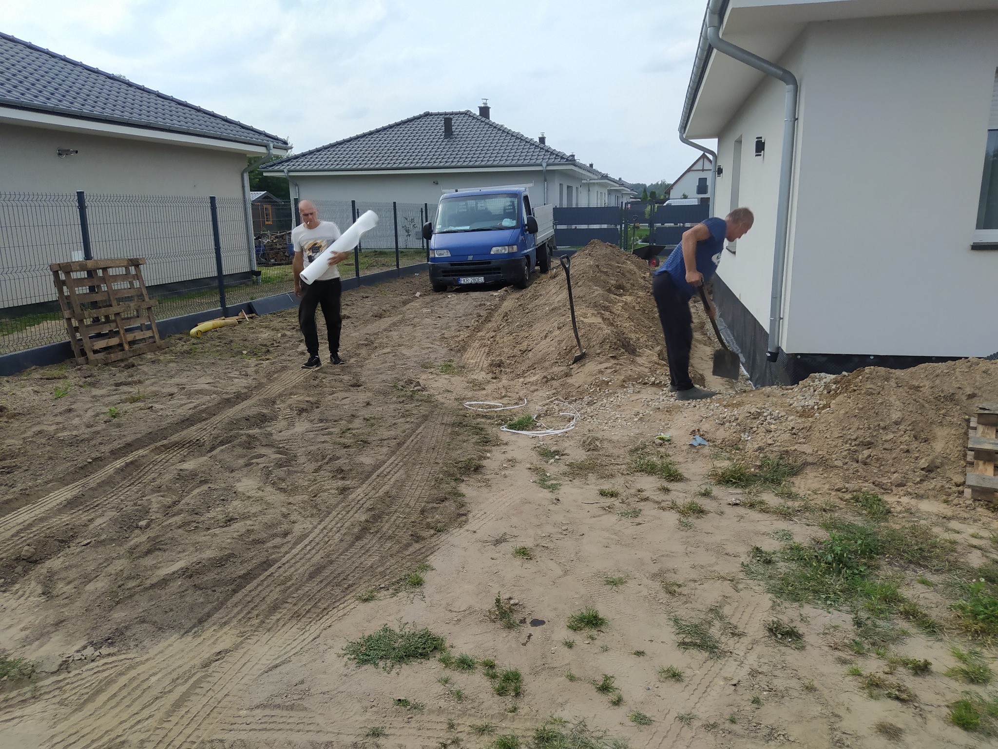 Prace ziemne przy nowym domu: mężczyzna kopie fundamenty szpadlem, drugi niesie izolację. Widoczny transport ziemi i przygotowany teren pod ogród.