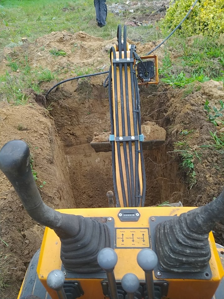 Widok z kabiny minikoparki na wykop w ziemi, z fragmentem osoby w tle. Żółta maszyna z czarnymi elementami sterującymi, otoczona zielenią.
