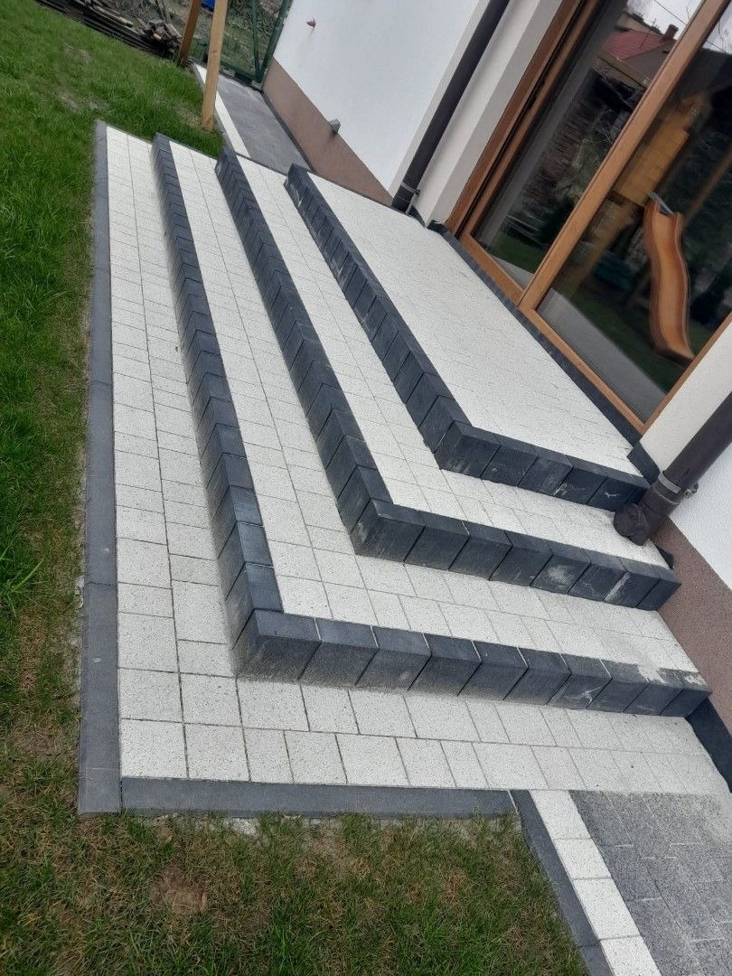 Schody z kostki brukowej w odcieniach szarości, prowadzące do domu z dużym oknem. Widoczne trawnik i fragment elewacji. Ułożenie kostki w geometryczny wzór.