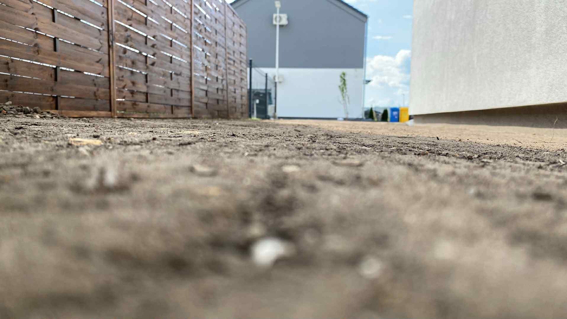 Przygotowany teren pod trawnik z rolki, widoczny drewniany płot i budynek w tle. Niska perspektywa podkreśla strukturę ziemi.
