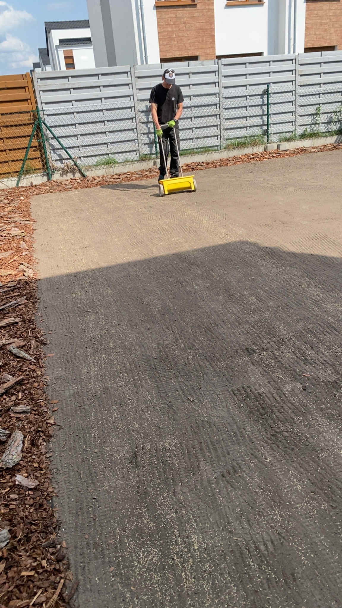 Pracownik z żółtym siewnikiem na przygotowanym podłożu, prawdopodobnie rozsiewa nasiona trawy. W tle ogrodzenie panelowe i budynek.