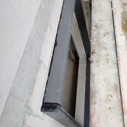 SLIMBUD Liubomyr Stefanko - Przygotowany otwór okienny z ościeżnicą zabezpieczoną czarną taśmą uszczelniającą, widok z góry na rusztowanie i surową ścianę z betonu komórkowego.