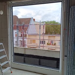 SLIMBUD Liubomyr Stefanko - Nowe okno PCV marki Schüco zamontowane w surowym stanie budynku, widok na zewnątrz z rusztowaniami i budynkami w tle, drabina oparta o ścianę.
