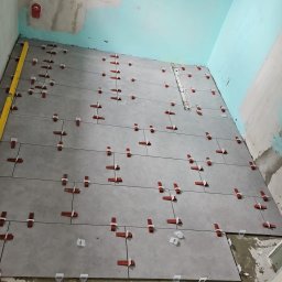 SLIMBUD Liubomyr Stefanko - Układanie szarych płytek podłogowych w łazience z użyciem poziomicy i czerwonych klipsów poziomujących, ściany w surowym stanie.