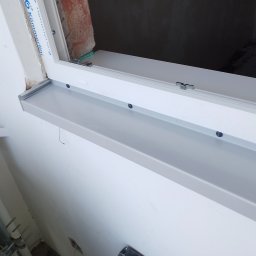 SLIMBUD Liubomyr Stefanko - Zamontowane okno PCV z szarym parapetem wewnętrznym, widoczne ślady po montażu na ścianie i nieotynkowane wnętrze wnęki okiennej.