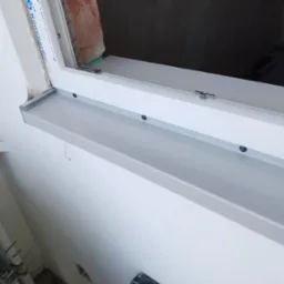 Zamontowane okno PCV z szarym parapetem wewnętrznym, widoczne ślady po montażu na ścianie i nieotynkowane wnętrze wnęki okiennej.