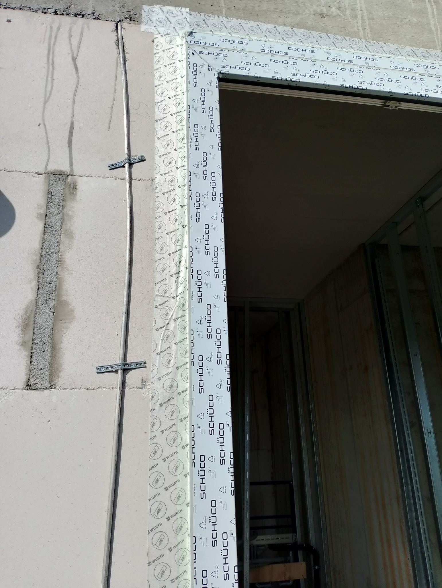 Surowy mur z zamontowaną ościeżnicą drzwiową marki Schüco, zabezpieczoną folią ochronną z nadrukiem logo, widoczne metalowe profile konstrukcyjne wewnątrz otworu.