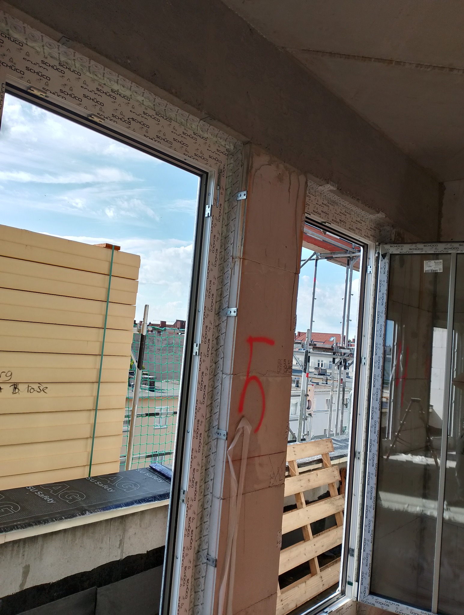 Nowe okno PCV marki Schüco zamontowane w surowym stanie budynku, widoczne elementy montażowe i izolacja termiczna na zewnątrz, zarys miasta w tle.