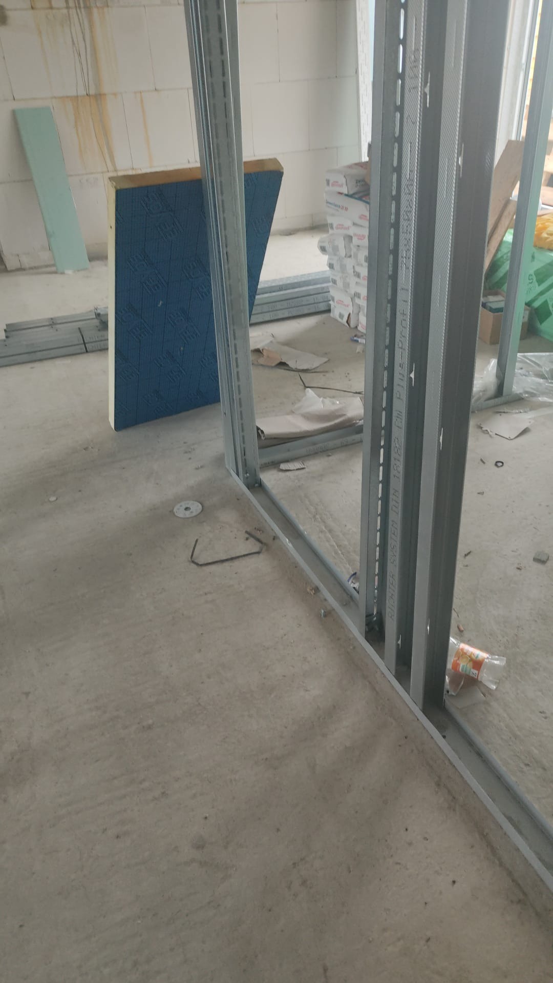 Konstrukcja szkieletowa ścian działowych z profili stalowych, widoczne płyty izolacyjne i materiały budowlane w tle, surowy stan wykończenia wnętrza.