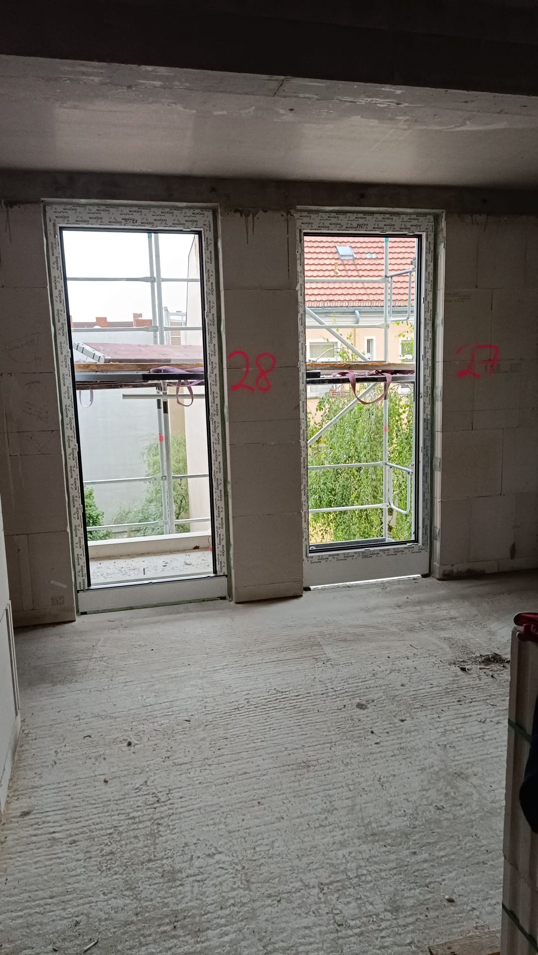 Surowy stan wnętrza z dwoma nowo zamontowanymi oknami PCV z widokiem na zewnątrz, widoczne rusztowania za oknami, ściany z betonu komórkowego z naniesionymi numerami.