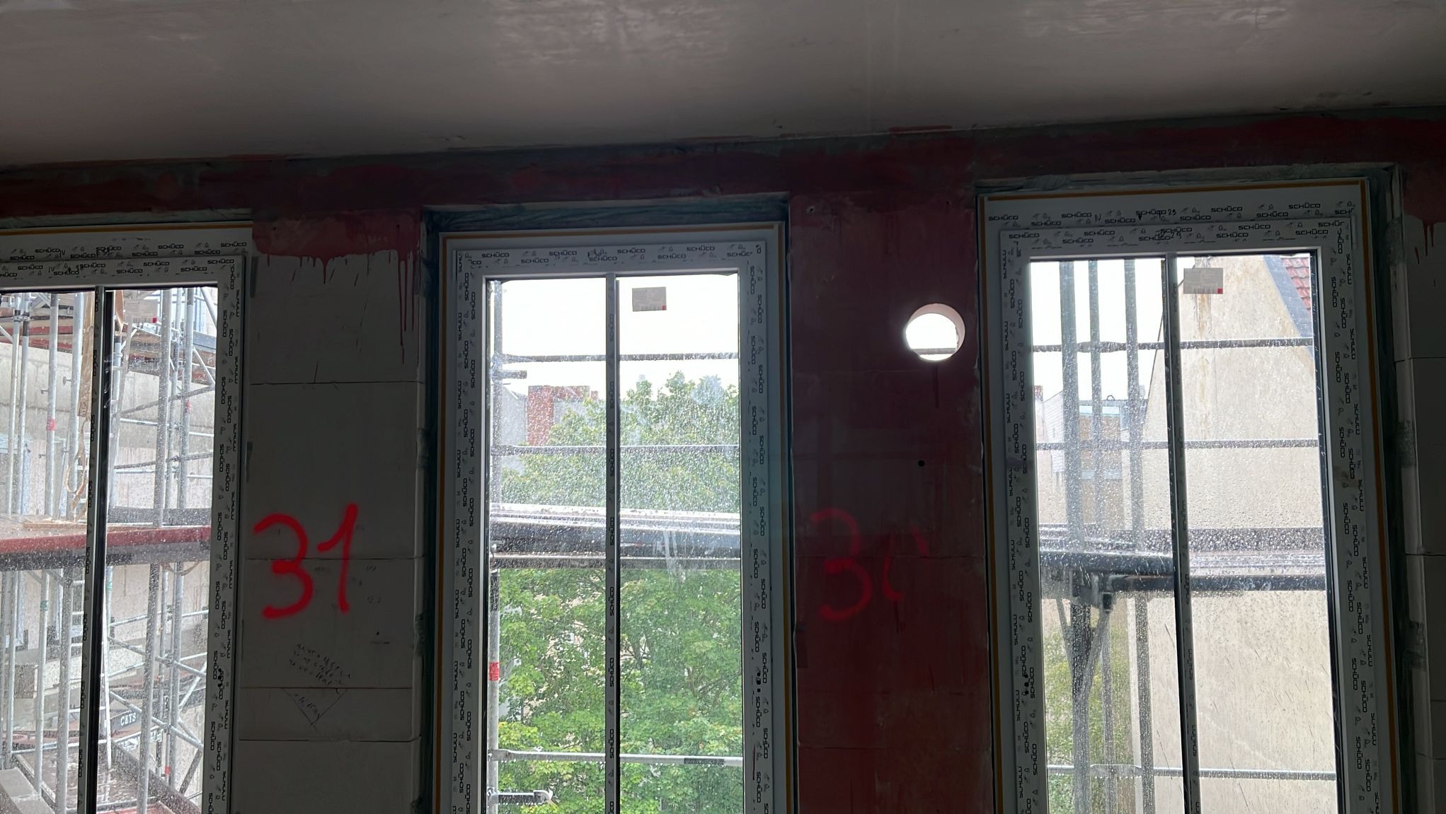Trzy okna PCV z widokiem na zewnątrz, zamontowane w surowym stanie, ściany w trakcie wykończenia, widoczne czerwone napisy '31' i '30' na ścianach.