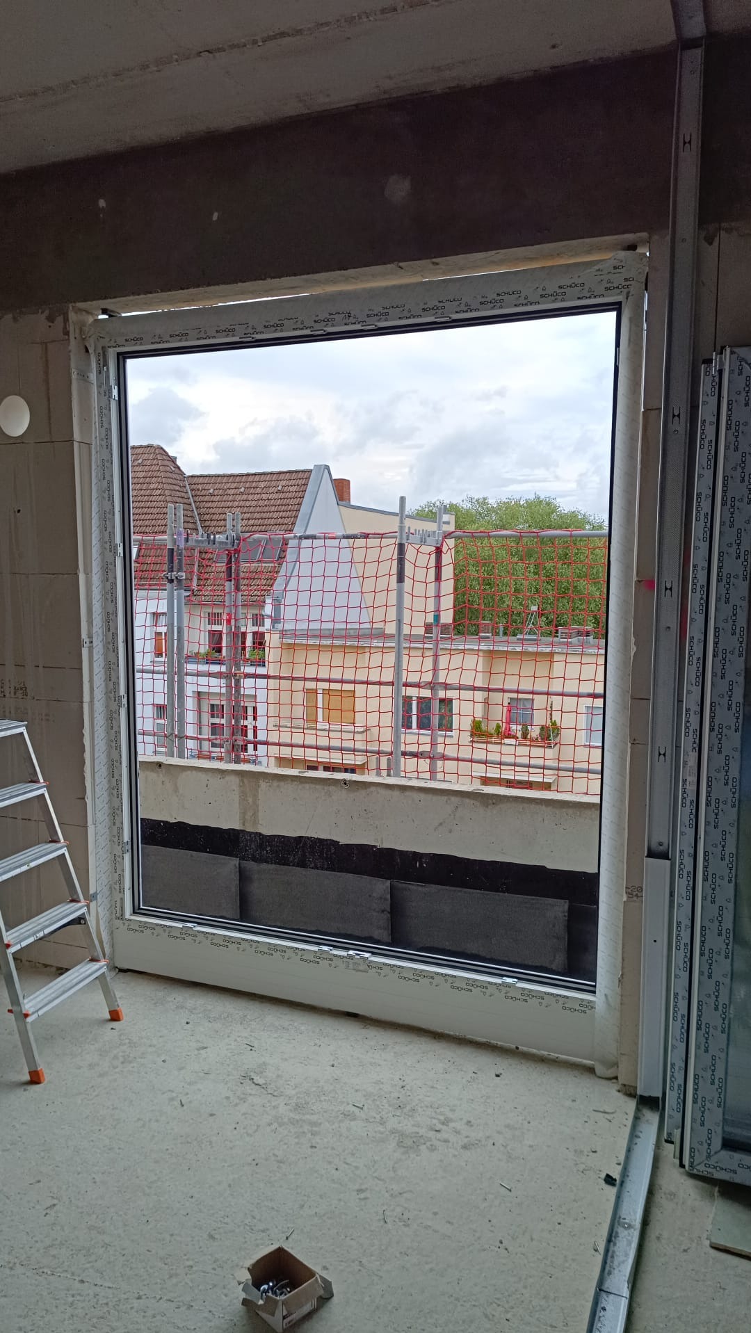 Nowe okno PCV marki Schüco zamontowane w surowym stanie budynku, widok na zewnątrz z rusztowaniami i budynkami w tle, drabina oparta o ścianę.