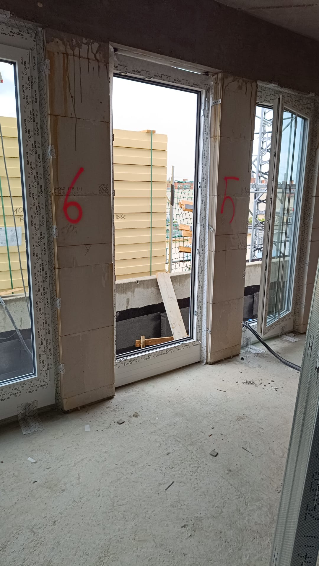 Wnętrze w trakcie montażu okien PCV, widoczne trzy otwory okienne z zamontowanymi ramami, ściany z surowego betonu z numerami naniesionymi czerwoną farbą, na zewnątrz widoczne płyty izolacyjne...