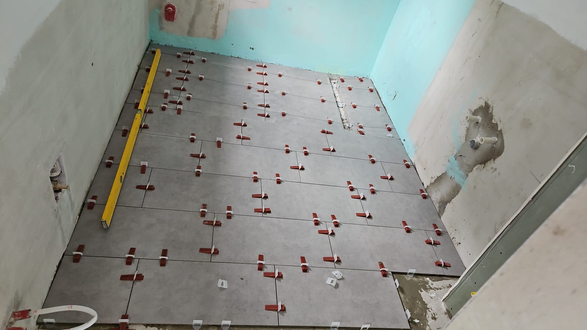 Układanie szarych płytek podłogowych w łazience z użyciem poziomicy i czerwonych klipsów poziomujących, ściany w surowym stanie.