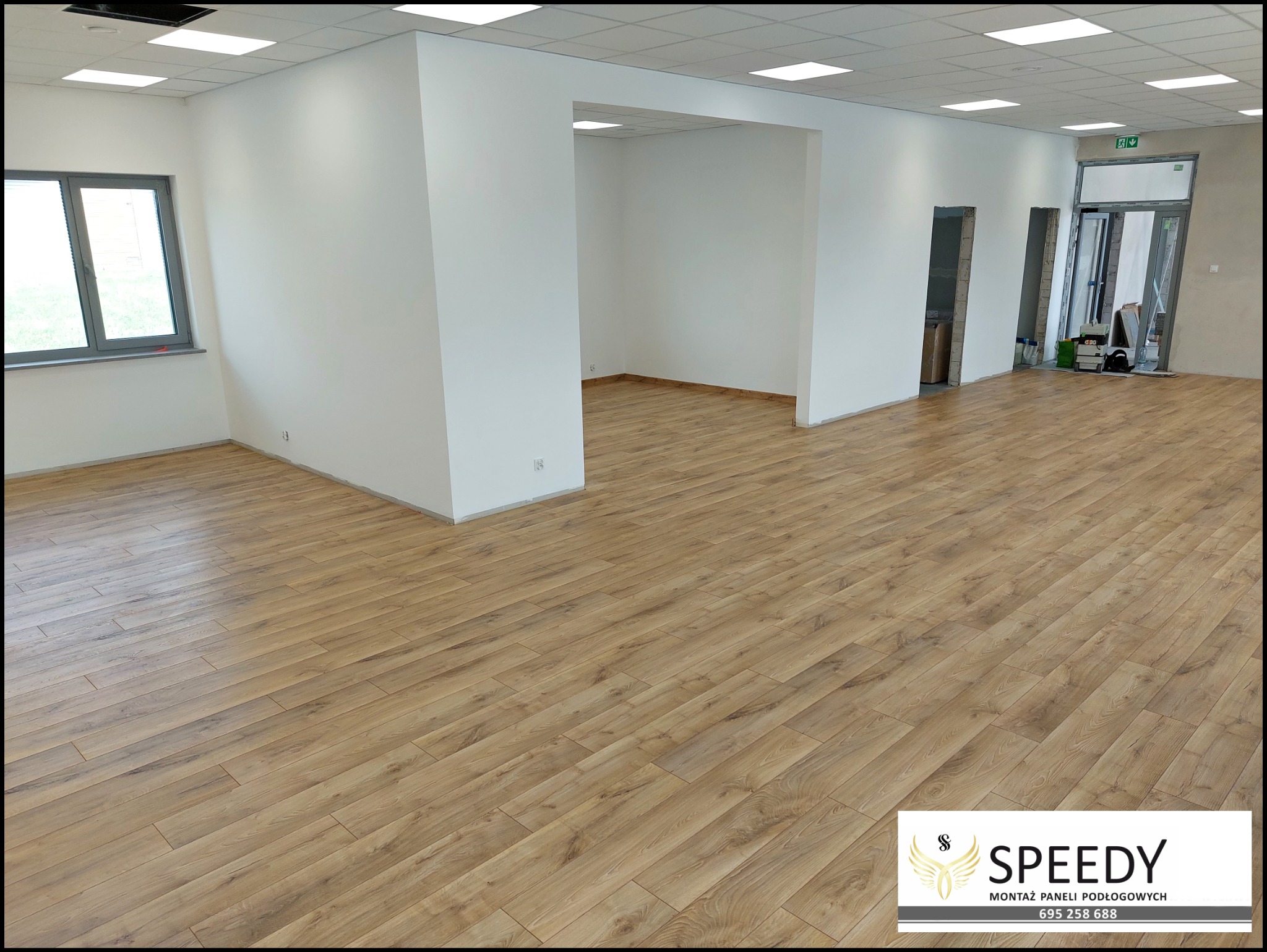 Jasne panele podłogowe ułożone diagonalnie w przestronnym, nowym biurze z białymi ścianami i oświetleniem sufitowym. Widoczne wejścia do innych pomieszczeń i logo firmy w rogu.