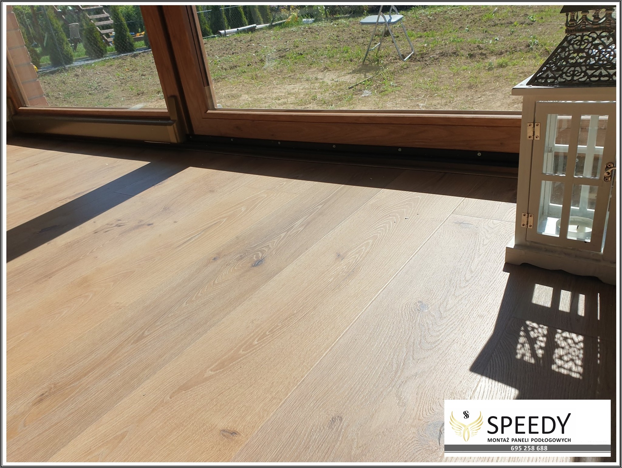 Jasne panele podłogowe z widocznym usłojeniem, ułożone wzdłuż okna tarasowego z drewnianą ramą, obok dekoracyjny lampion.