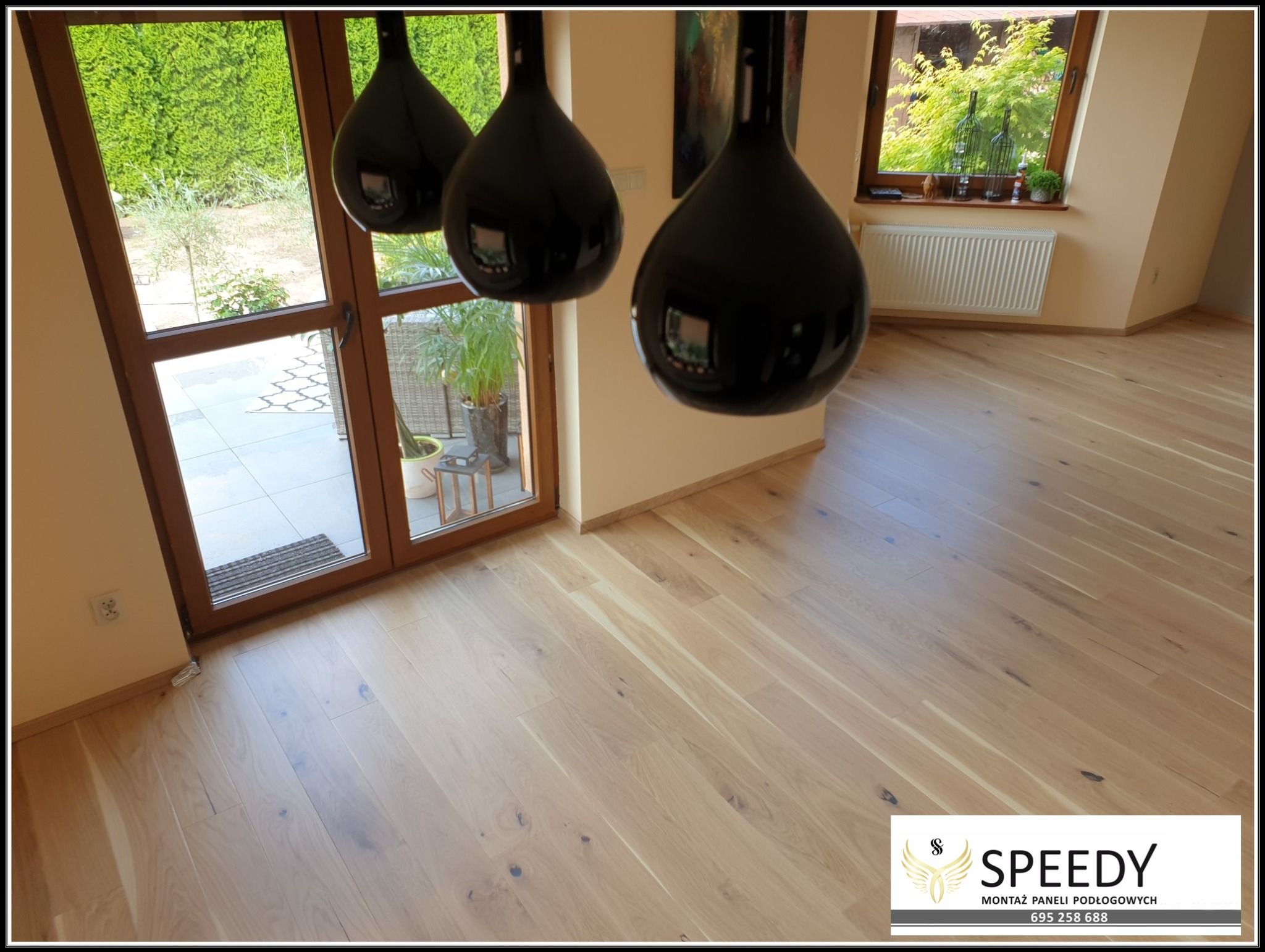 Jasne panele podłogowe ułożone w salonie z widokiem na ogród przez przeszklone drzwi balkonowe, wiszące czarne lampy kuliste w tle.