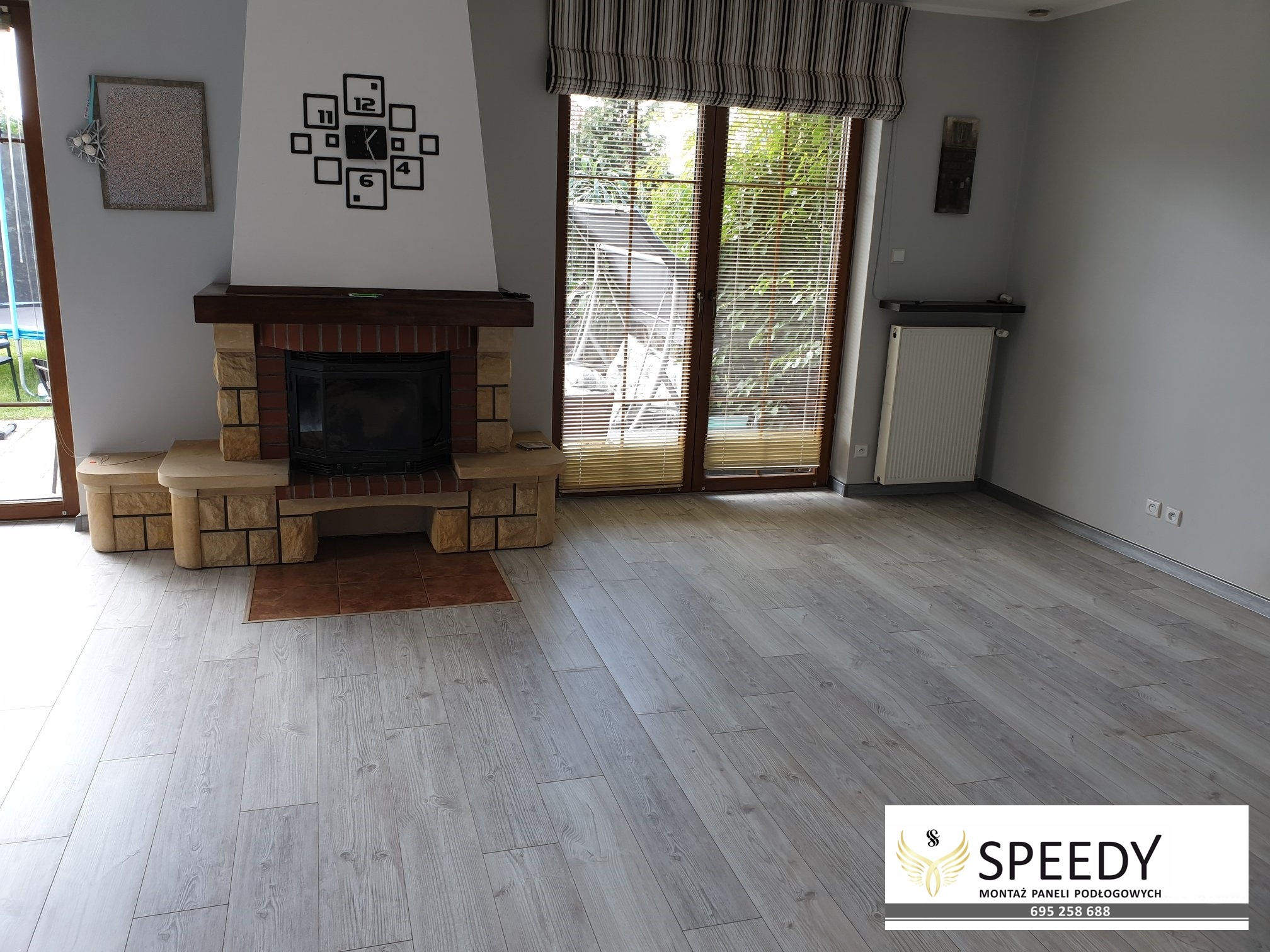 Jasnoszara podłoga z paneli ułożona w salonie z kominkiem, widoczne okno z żaluzjami i dekoracyjny zegar na ścianie.