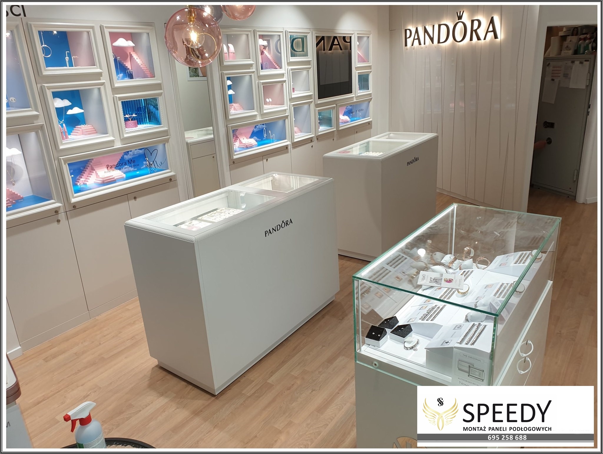 Wnętrze sklepu Pandora z drewnianą podłogą, białymi meblami ekspozycyjnymi i dekoracyjnymi gablotami ściennymi z motywami chmur i schodów. W tle widoczne logo Pandora. W rogu kadru butelka ze...