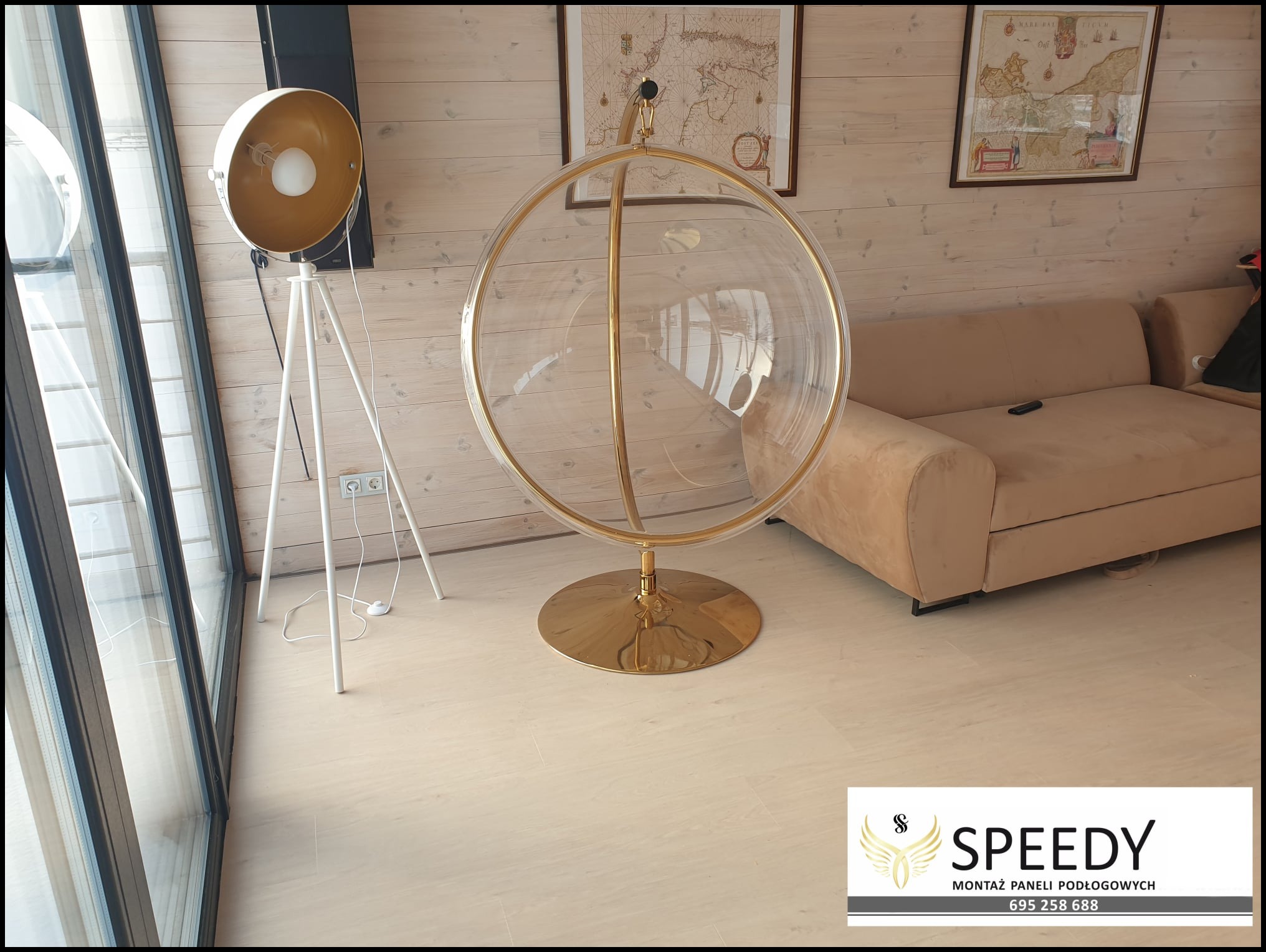 Jasne panele podłogowe w salonie z designerskim fotelem-kulą na złotej podstawie i lampą na trójnogu, widoczne mapy na ścianie i fragment sofy.