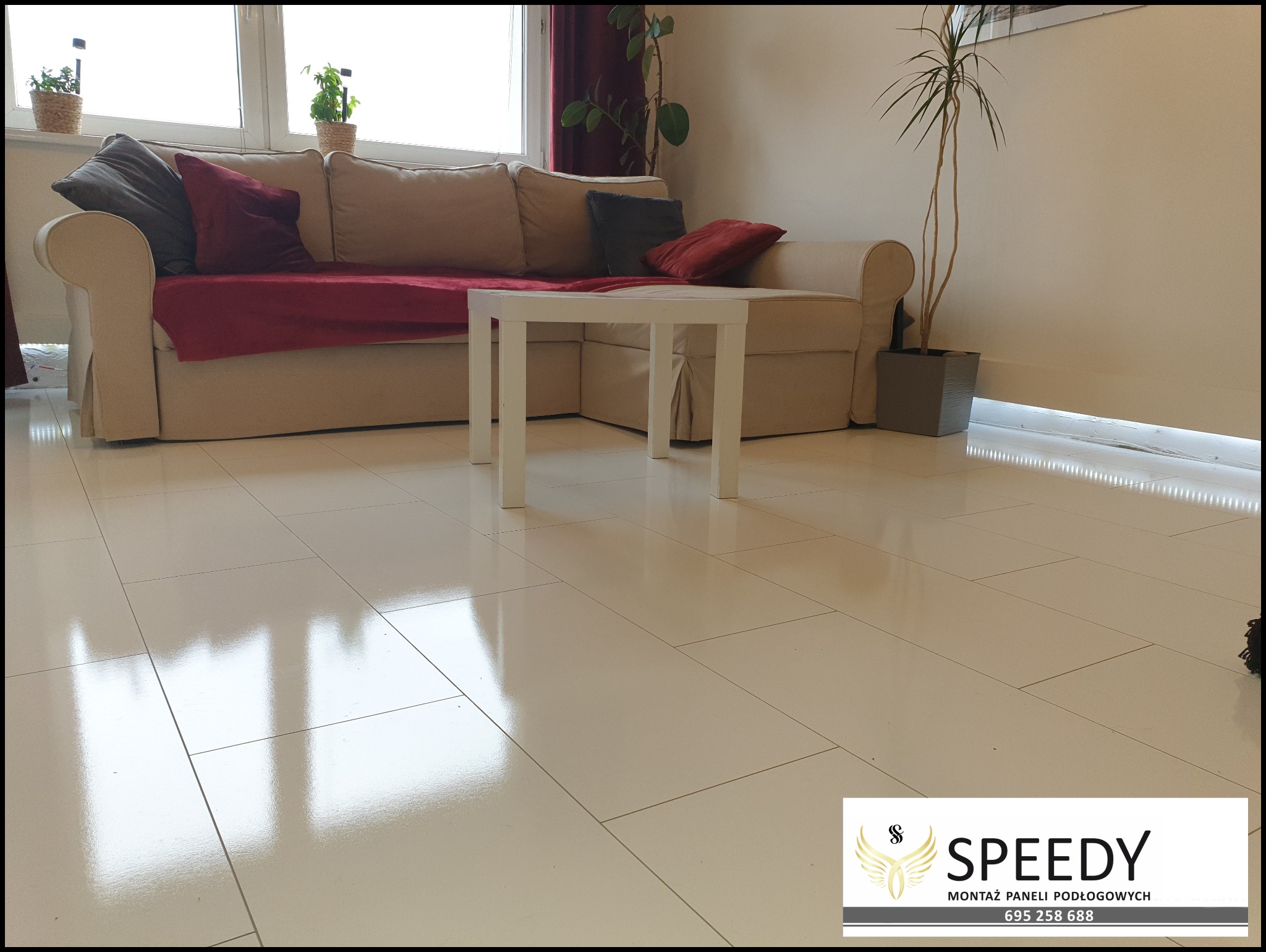 Jasne, błyszczące panele podłogowe ułożone w salonie z beżową kanapą, białym stolikiem i roślinami doniczkowymi, oświetlone listwą LED przy podłodze.