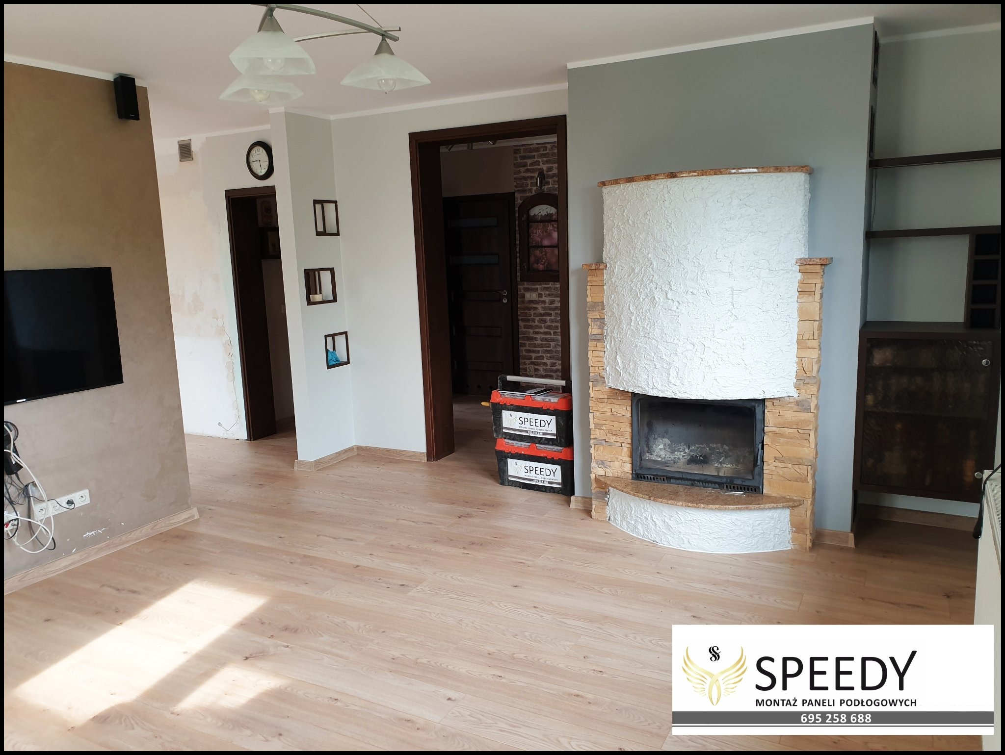 Jasne panele podłogowe w salonie z kominkiem, widoczne narzędzia w skrzynkach firmy Speedy, ściany w różnych odcieniach szarości i beżu.