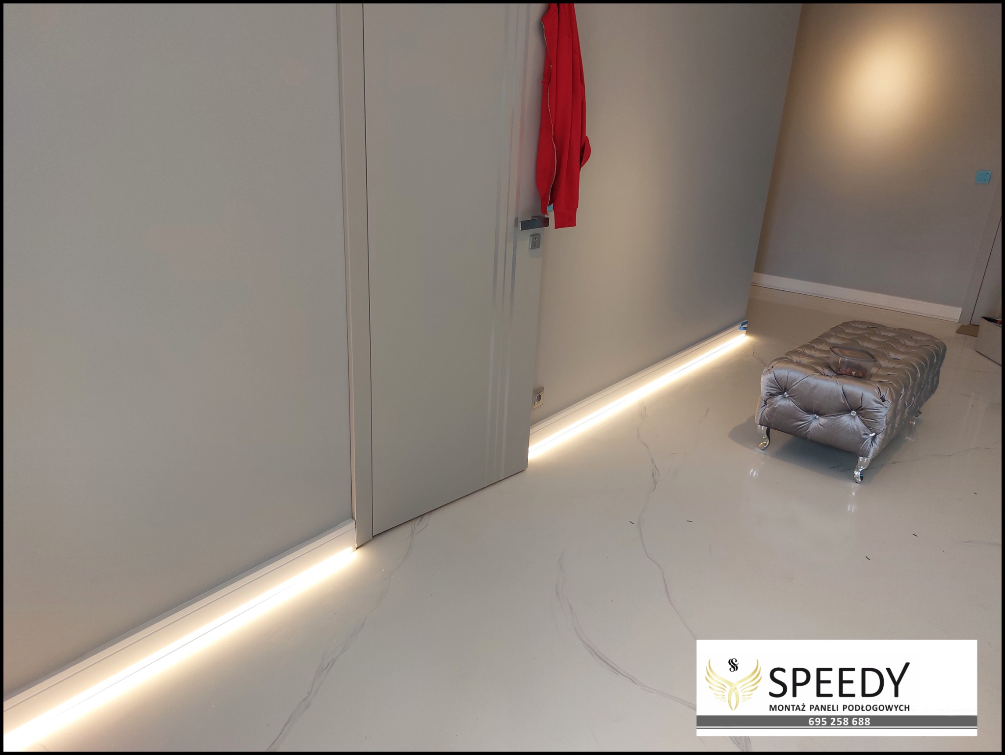 Biała marmurowa podłoga z listwami przypodłogowymi LED, szara ściana, drzwi z czerwoną bluzą, pufa w tle.