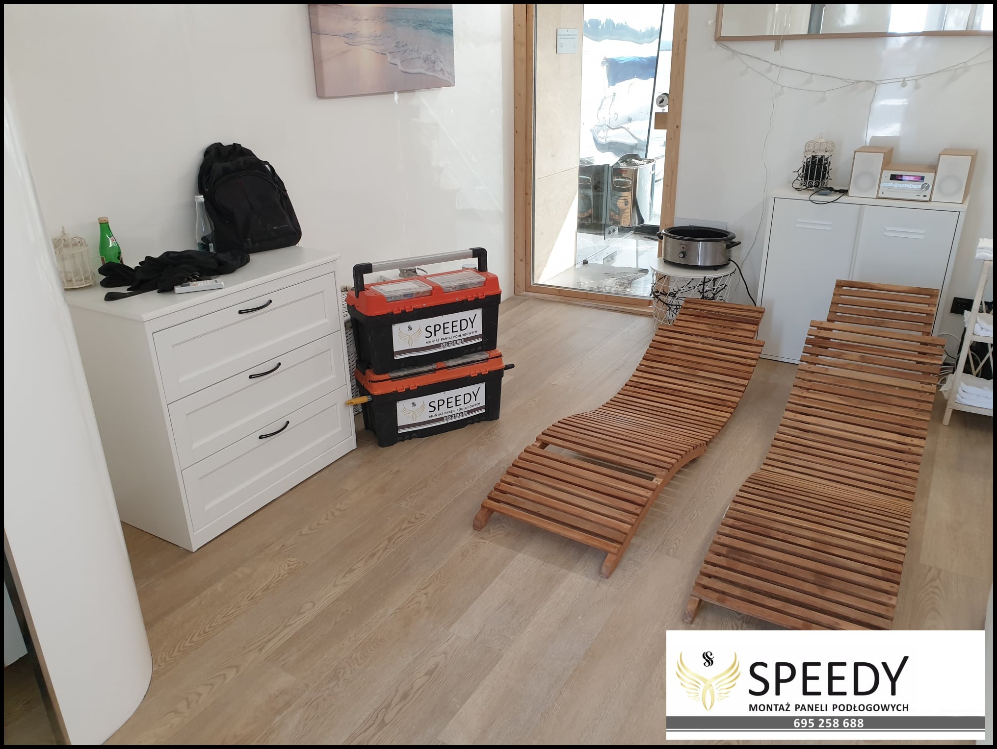 Jasne panele podłogowe z widocznymi narzędziami w firmowych skrzynkach i dwoma drewnianymi leżakami w pomieszczeniu z białymi meblami.