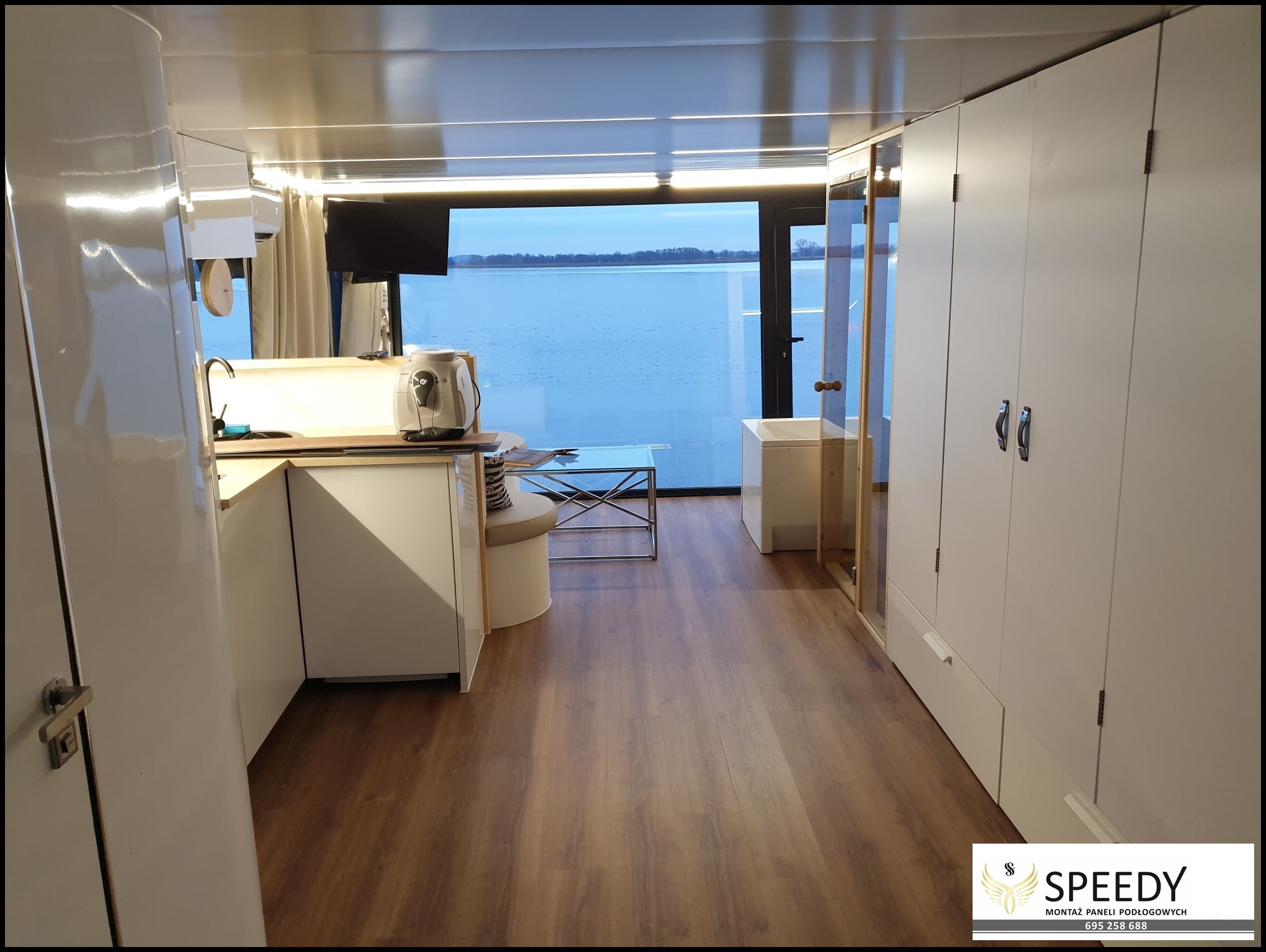 Wnętrze pływającego domu z panelami podłogowymi w odcieniach brązu, widok na jezioro przez panoramiczne okno.