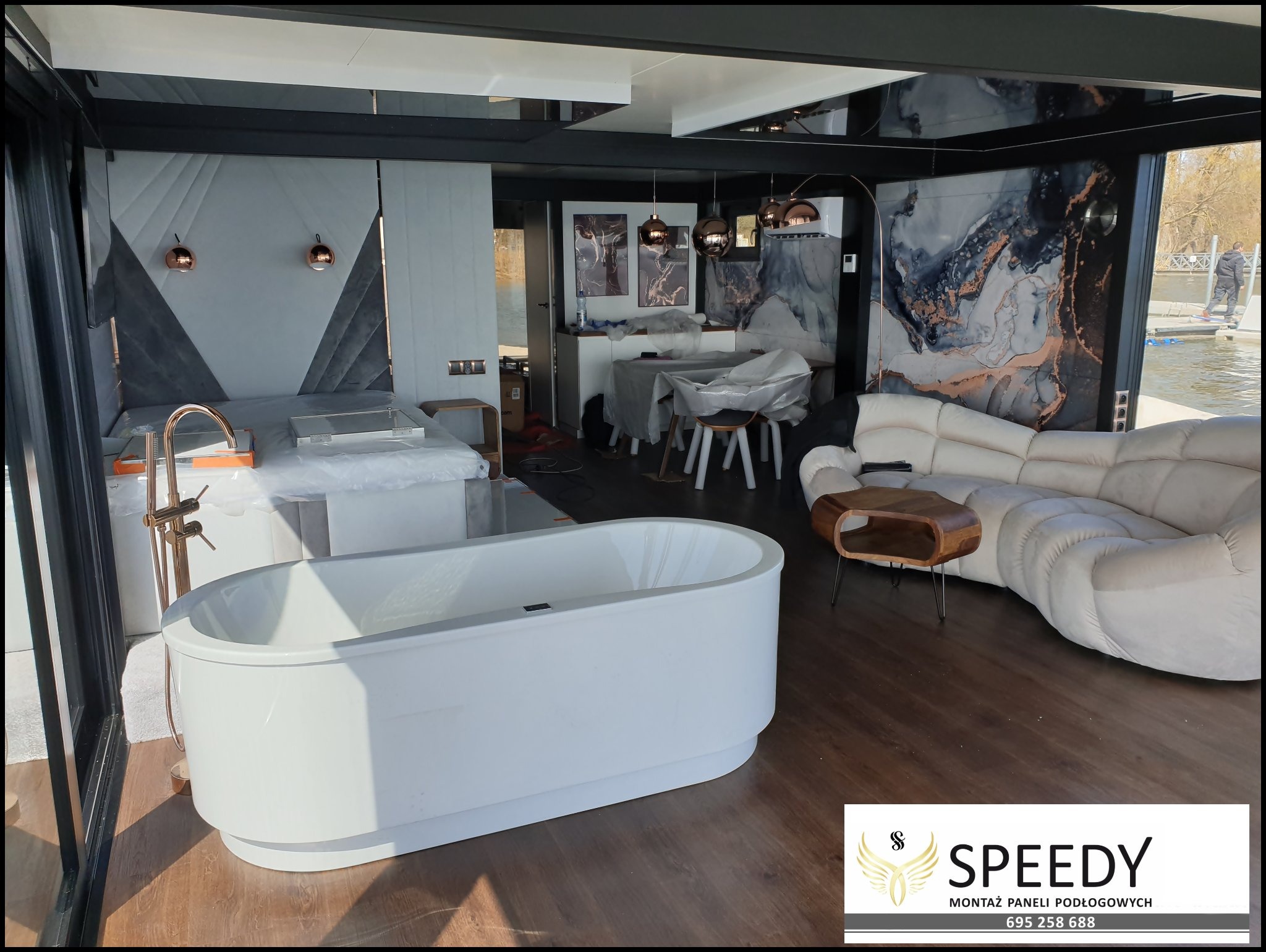 Nowoczesne wnętrze domu na wodzie z ciemnymi panelami podłogowymi, białą wanną wolnostojącą, falującą sofą i widokiem na rzekę.