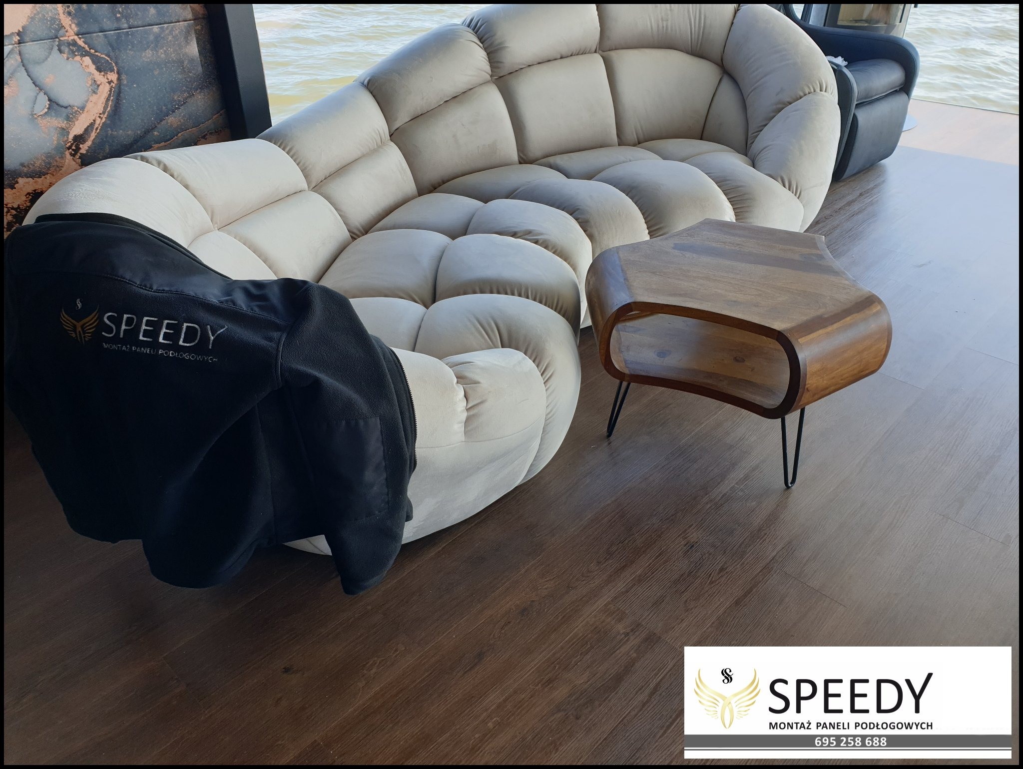 Koszalin: Nowoczesne panele podłogowe w odcieniach brązu, luksusowa sofa w kształcie muszli i stolik kawowy na tle wody, z firmową bluzą z logo.