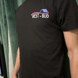 Kajot Design Graphic - Czarna koszulka z nadrukiem BEST-BUD z motywem domu w kolorach różowym i niebieskim, na tle zielonej zasłony.