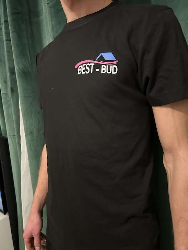 Czarna koszulka z nadrukiem BEST-BUD z motywem domu w kolorach różowym i niebieskim, na tle zielonej zasłony.