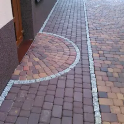 Ułożona z kostki brukowej ścieżka przed domem, z półkolistym wzorem przy wejściu, obrzeża z jasnoszarego kamienia.