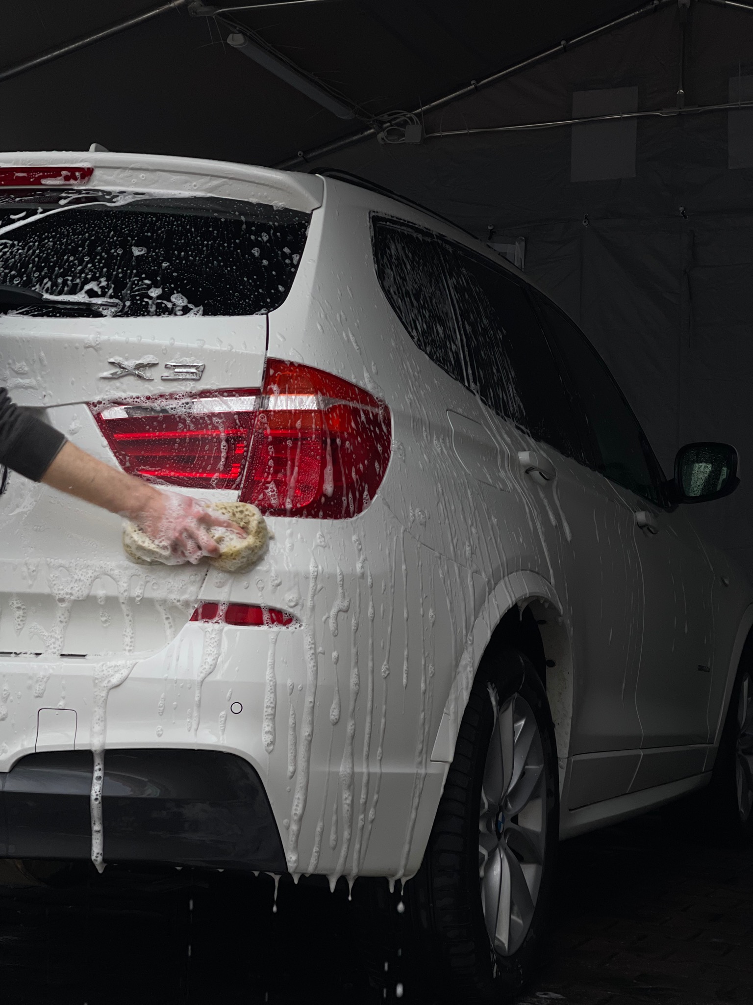 Mycie tylnej lampy białego BMW X3 gąbką, piana spływa po karoserii, widoczna ręka pracownika.