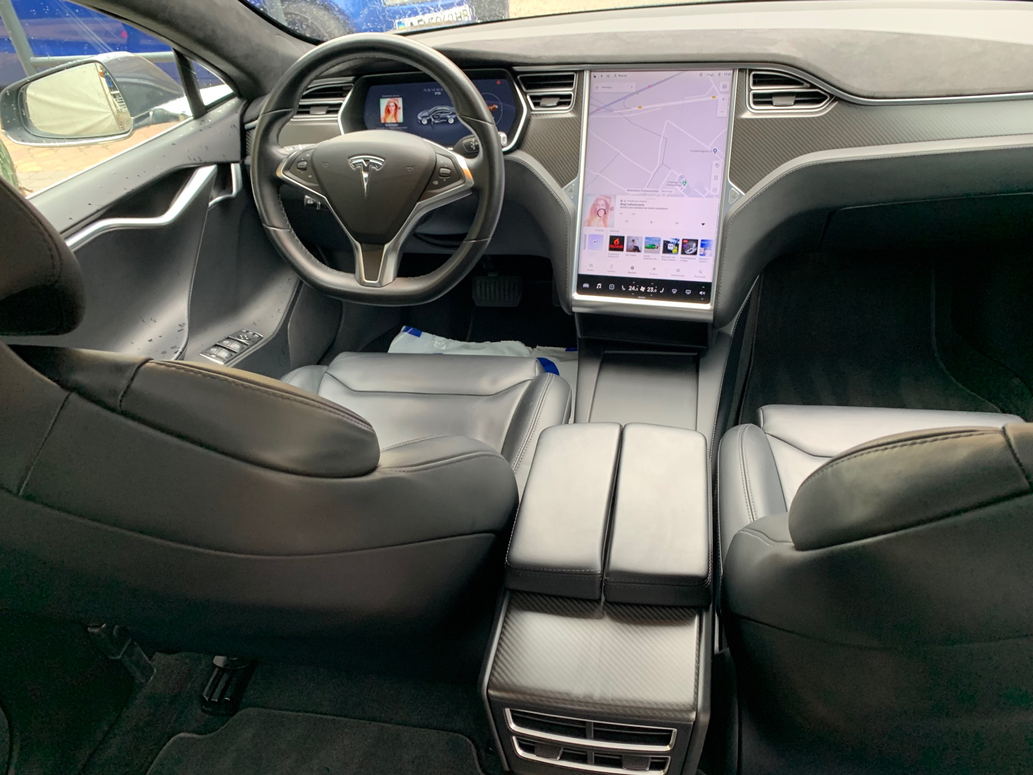 Wnętrze samochodu Tesla z czarną skórzaną tapicerką i karbonowymi elementami deski rozdzielczej, widoczny duży ekran nawigacji i kierownica z logo Tesla.