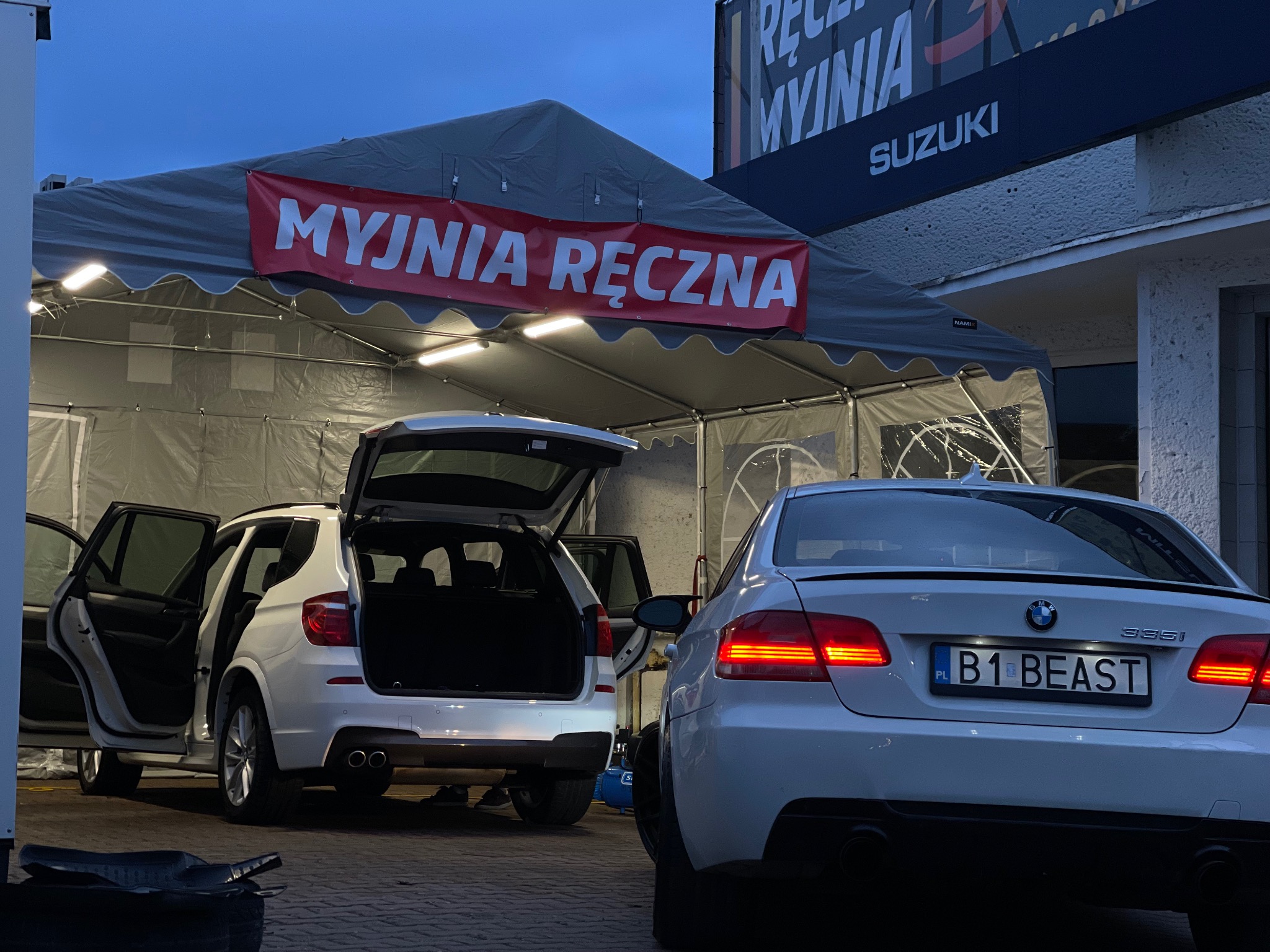 Dwa białe samochody BMW, X3 z otwartymi drzwiami i bagażnikiem oraz 335i z tablicą rejestracyjną B1 BEAST, stoją pod namiotem z napisem 'Myjnia Ręczna', obok budynku z szyldem reklamowym.