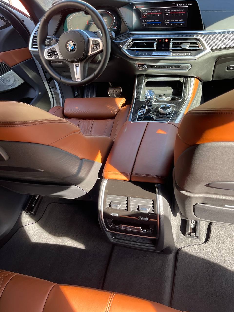 Wnętrze samochodu BMW X6 z brązową skórzaną tapicerką, widok z tylnych siedzeń na deskę rozdzielczą i kierownicę, oświetlone popołudniowym słońcem.