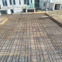 Firma Budowlana Marbis - Siatka zbrojeniowa na fundamencie budynku w trakcie budowy, widok z góry. Elementy szalunku widoczne w tle, surowy stan.