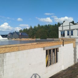 Firma Budowlana Marbis - Budowa domu z białych bloczków: ściany parteru z oknem, strop zalany betonem. W tle inne domy w stanie surowym, las i błękitne niebo z chmurami.