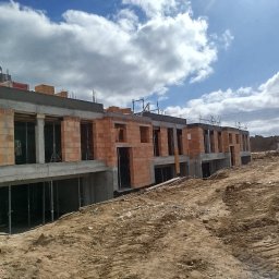 Firma Budowlana Marbis - Budowa szeregowców z cegły ceramicznej. Widoczny murarz na dachu, elementy konstrukcyjne i teren budowy. Pochmurne niebo w tle. Ujęcie z dołu.