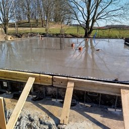 Firma Budowlana Marbis - Zalany beton na fundamencie z wystającymi prętami zbrojeniowymi i elementami instalacji. Widoczne szalunki i drewniane podpory. W tle drzewa i trawa.