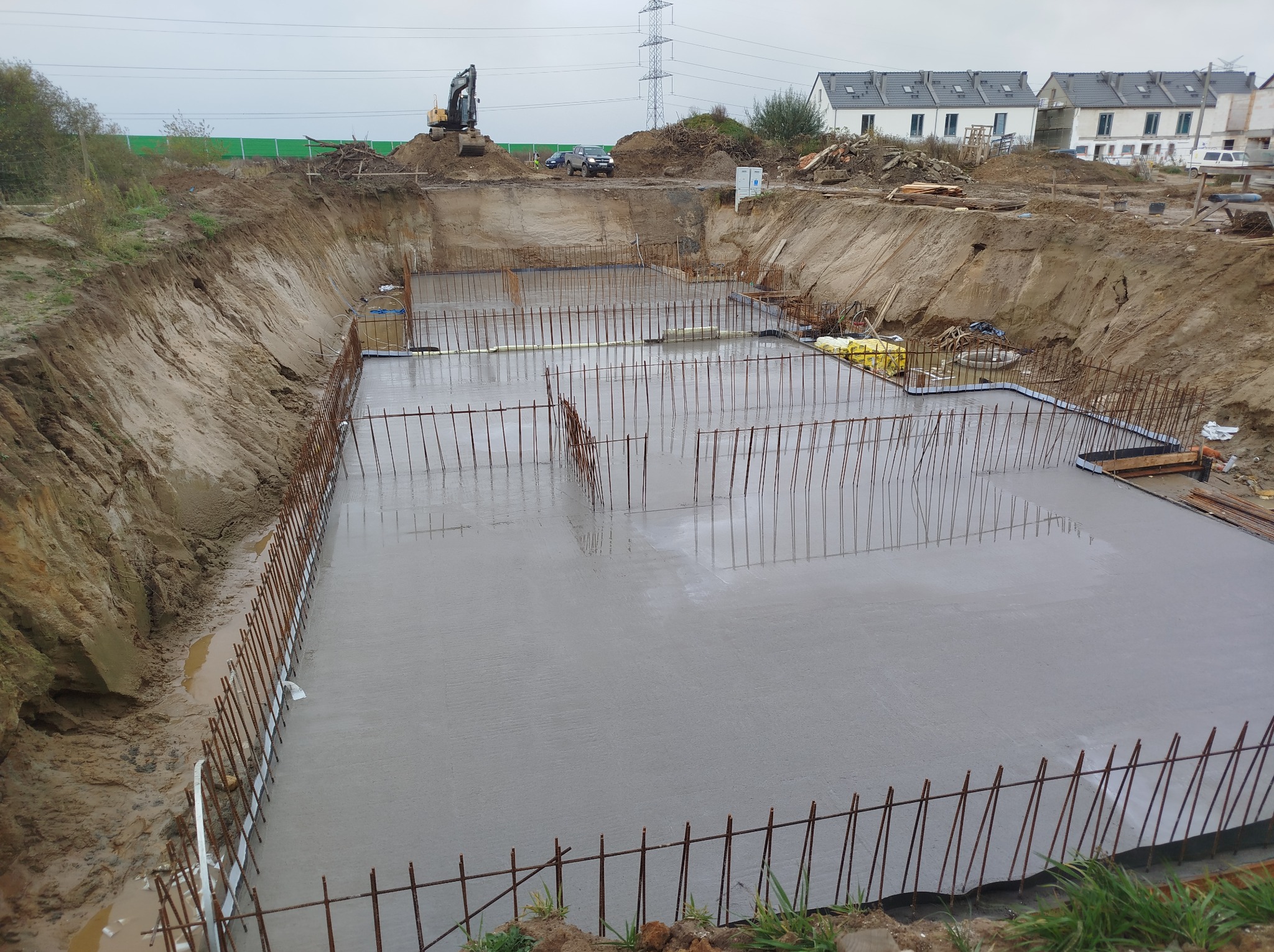 Zalany betonem fundament z wystającą stalą zbrojeniową w wykopie. W tle budynki mieszkalne i koparka. Widok z góry na plac budowy.