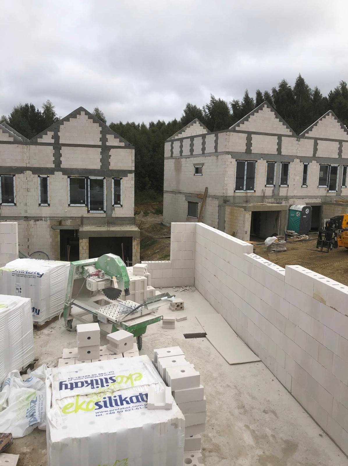 Budowa domów z białych bloków silikatowych, widoczna piła do cięcia bloczków, palety z materiałem budowlanym. Domy w stanie surowym otwartym, tło stanowią drzewa.
