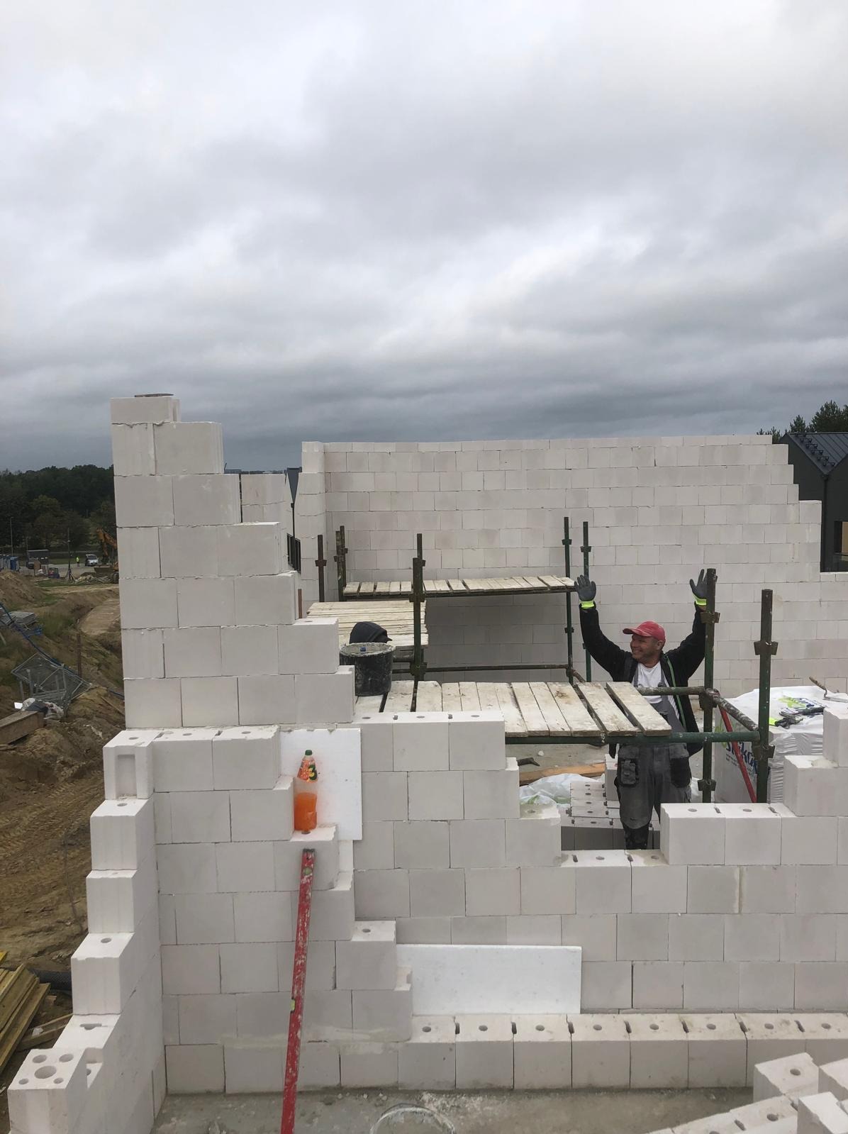Wznoszenie muru z białych bloczków, widoczny murarz na rusztowaniu z podniesionymi rękami, butelka z napojem na murze. Budowa domu w toku, pochmurne niebo w tle.