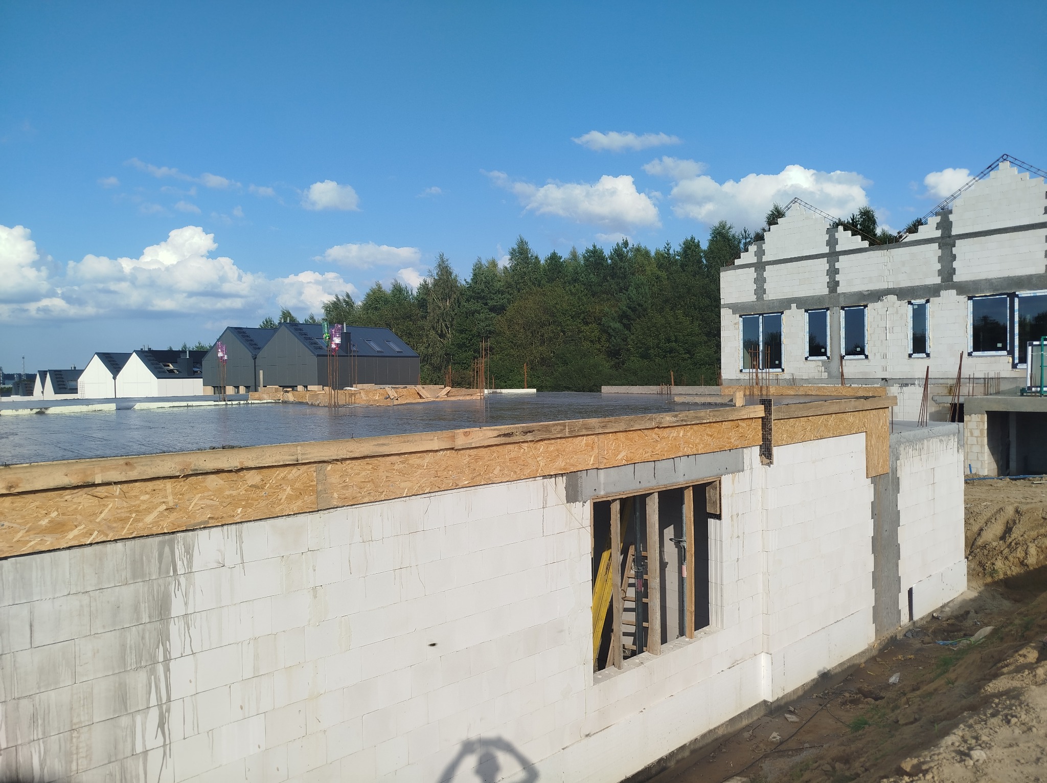 Budowa domu z betonu komórkowego: ściany parteru z oknami, strop zalany betonem. W tle inne budynki w stanie surowym i las. Błękitne niebo z chmurami.