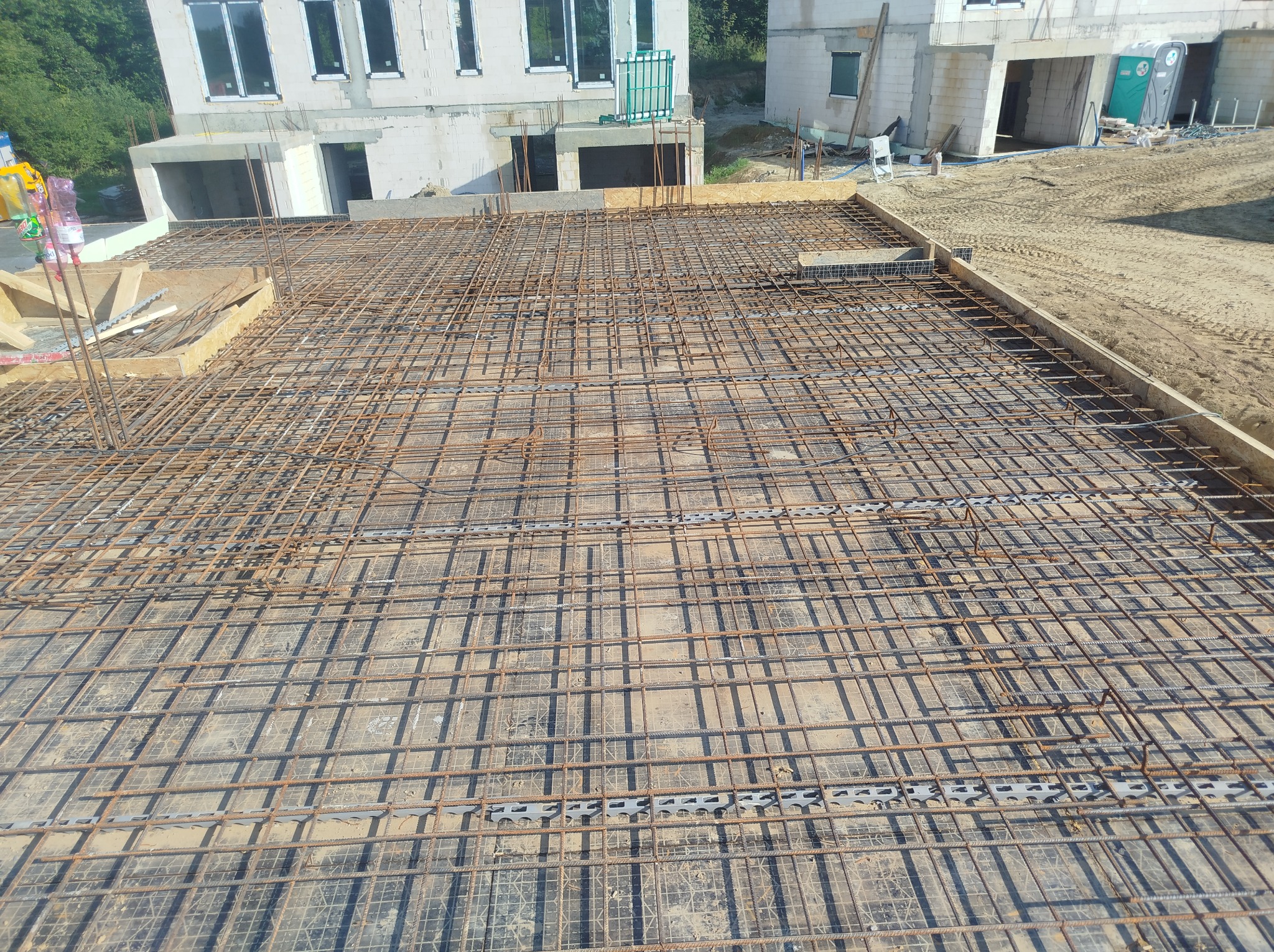 Siatka zbrojeniowa na fundamencie budynku w trakcie budowy, widok z góry. Elementy szalunku widoczne w tle, surowy stan.